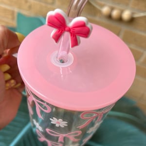 May include: A pink reusable tumbler with a clear lid and a pink bow straw topper. The tumbler has a clear glass body with a pink bow and daisy design.