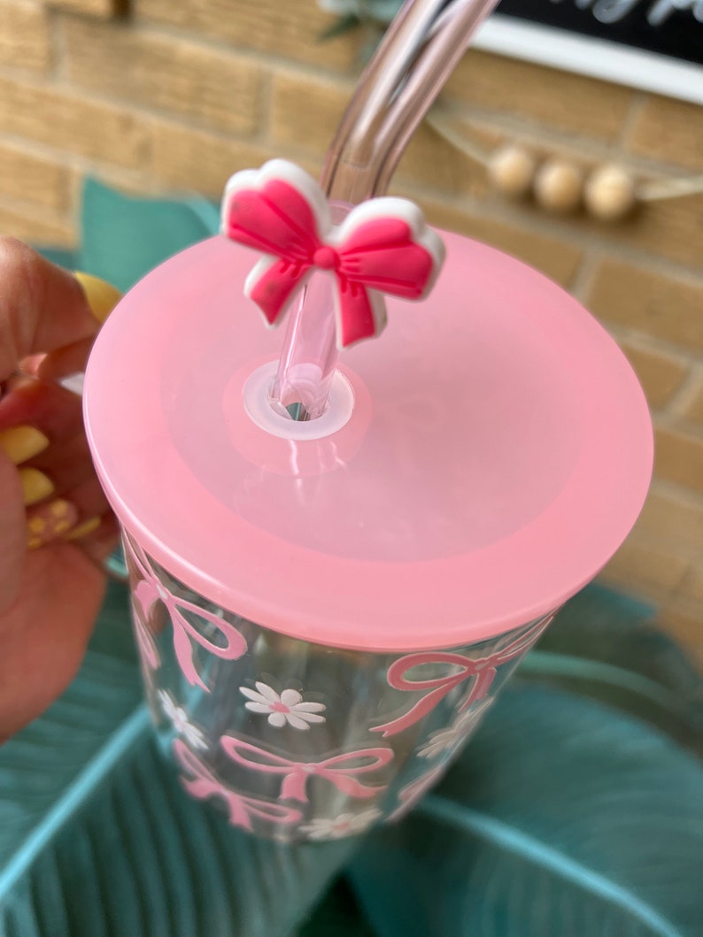 May include: A pink reusable tumbler with a clear lid and a pink bow straw topper. The tumbler has a clear glass body with a pink bow and daisy design.