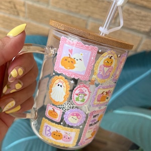 May include: A clear glass mug with a wooden lid and a white ghost-shaped straw topper. The mug is decorated with Halloween-themed illustrations in ornate frames, including pumpkins and ghosts. The mug is held by a person with yellow nail polish.