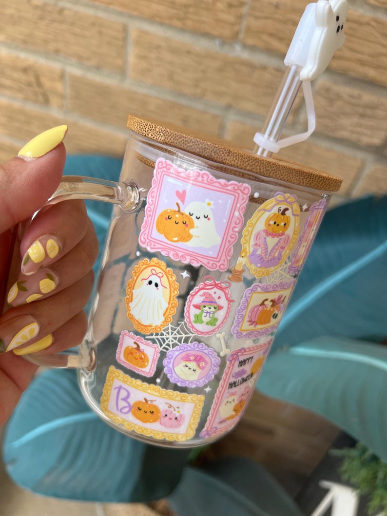 May include: A clear glass mug with a wooden lid and a white ghost-shaped straw topper. The mug is decorated with Halloween-themed illustrations in ornate frames, including pumpkins and ghosts. The mug is held by a person with yellow nail polish.