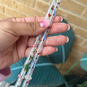Op de afbeelding: Twee doorzichtige plastic rietjes versierd met kleurrijke kerstverlichting, ornamenten en hulst. De rietjes worden vastgehouden in een hand met een roze en witte manicure. De rietjes zijn ontworpen voor feestelijke drankjes.