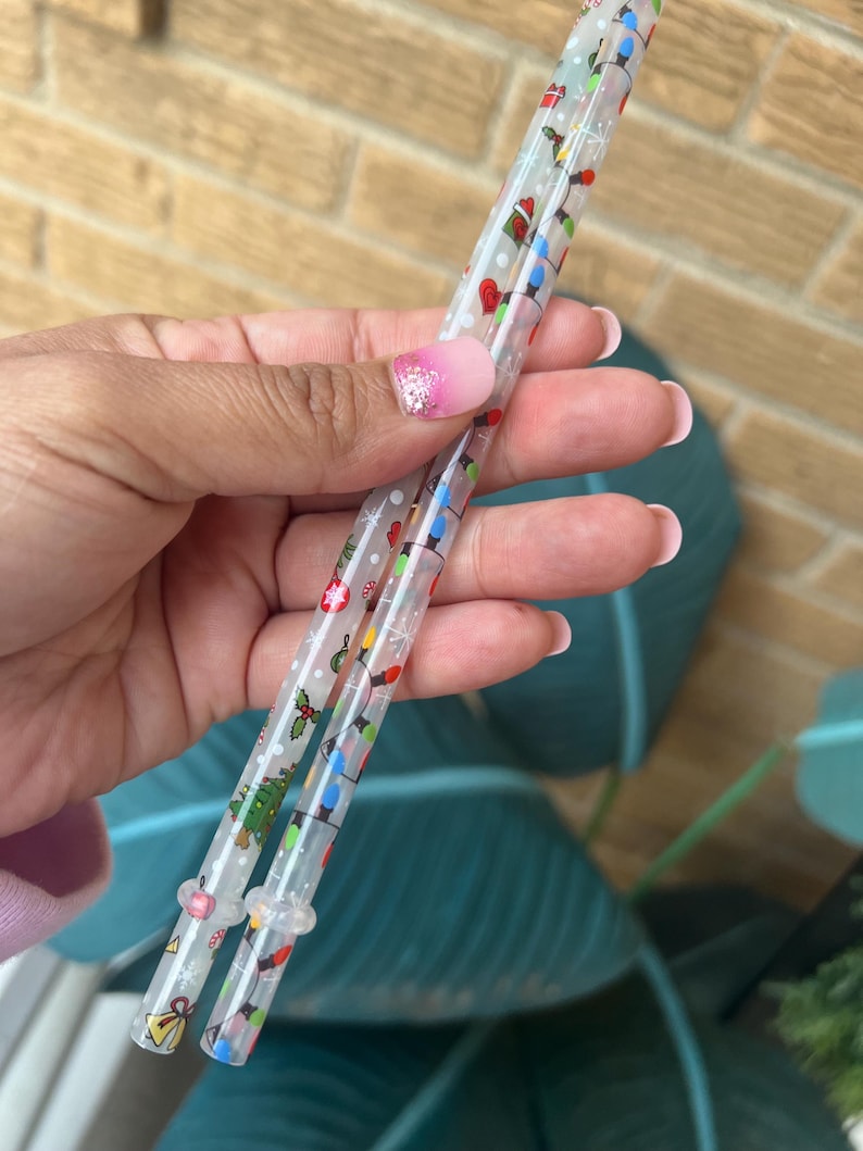 Op de afbeelding: Twee doorzichtige plastic rietjes versierd met kleurrijke kerstverlichting, ornamenten en hulst. De rietjes worden vastgehouden in een hand met een roze en witte manicure. De rietjes zijn ontworpen voor feestelijke drankjes.