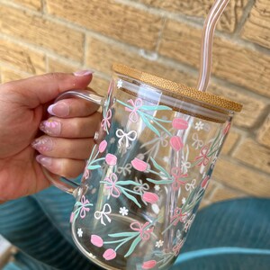 May include: A clear glass mug with a bamboo lid and a pink and green floral design. The mug has a handle and comes with a clear glass straw.