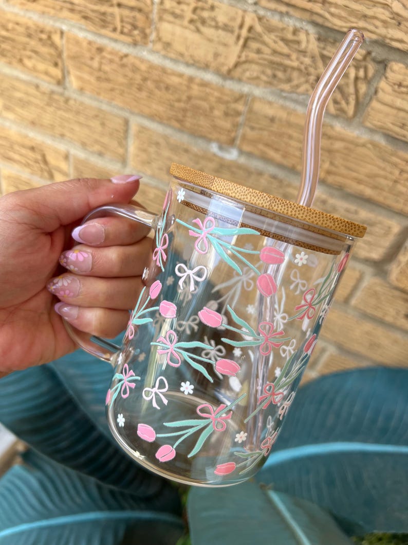 May include: A clear glass mug with a bamboo lid and a pink and green floral design. The mug has a handle and comes with a clear glass straw.
