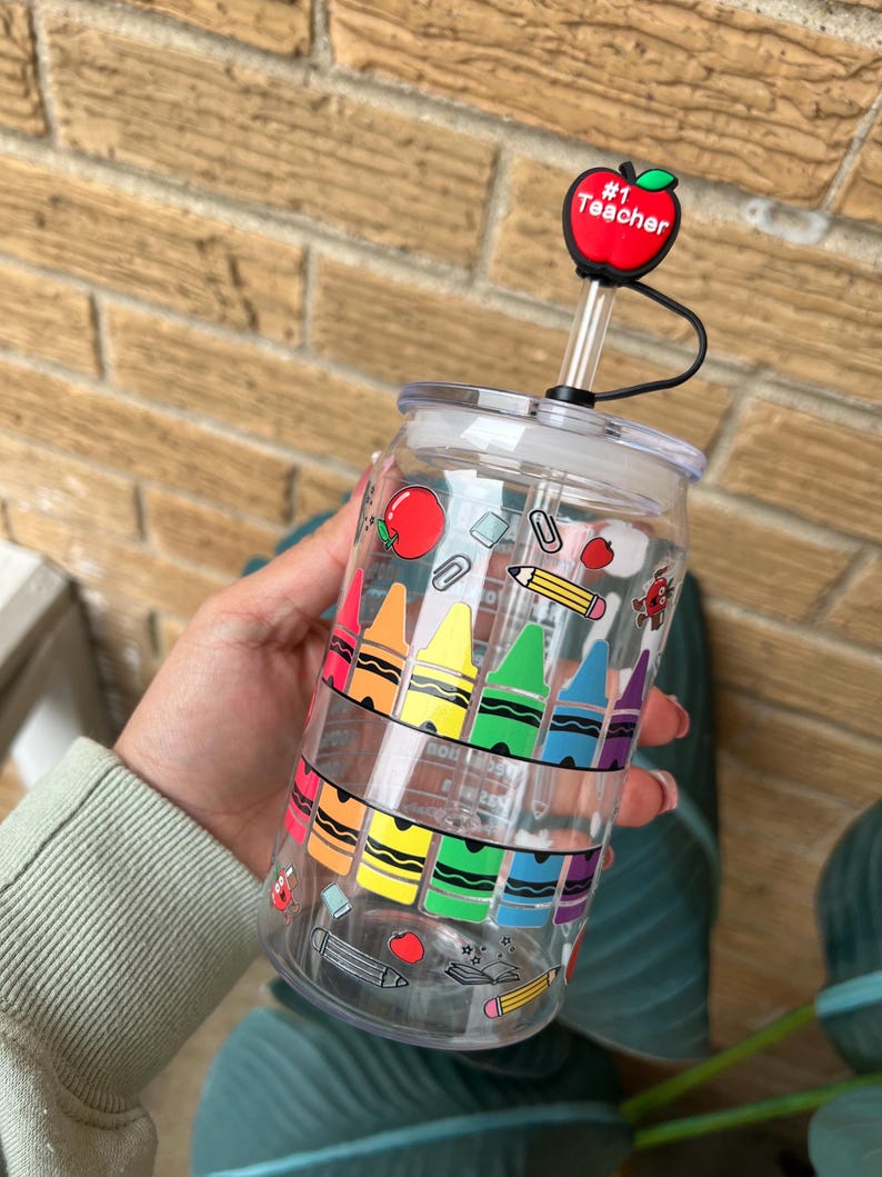 May include: Clear plastic reusable water bottle with a rainbow of crayons, pencils, and other school supplies printed on the side. The bottle has a red apple lid with the text "#1 Teacher" on it.