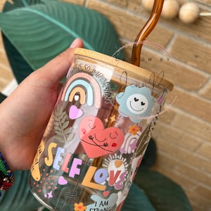 May include: A glass tumbler with a bamboo lid and a brown glass straw. The tumbler is decorated with colorful stickers that say "Self Love", "I am strong", and "Self care is so important".