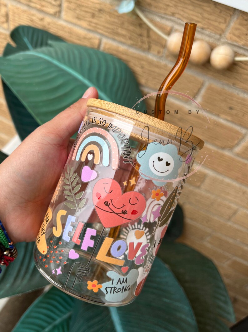 May include: A glass tumbler with a bamboo lid and a brown glass straw. The tumbler is decorated with colorful stickers that say "Self Love", "I am strong", and "Self care is so important".