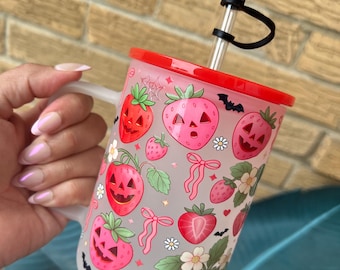 May include: A clear glass tumbler with a red lid and a black bat-shaped straw topper. The tumbler is decorated with pink strawberries with jack-o'-lantern faces, bows, and flowers. The handle is white.