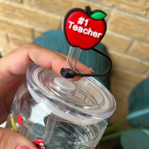 May include: A clear plastic water bottle with a red apple lid that says "#1 Teacher". The bottle has a straw and is decorated with school supplies like pencils, rulers, and apples.