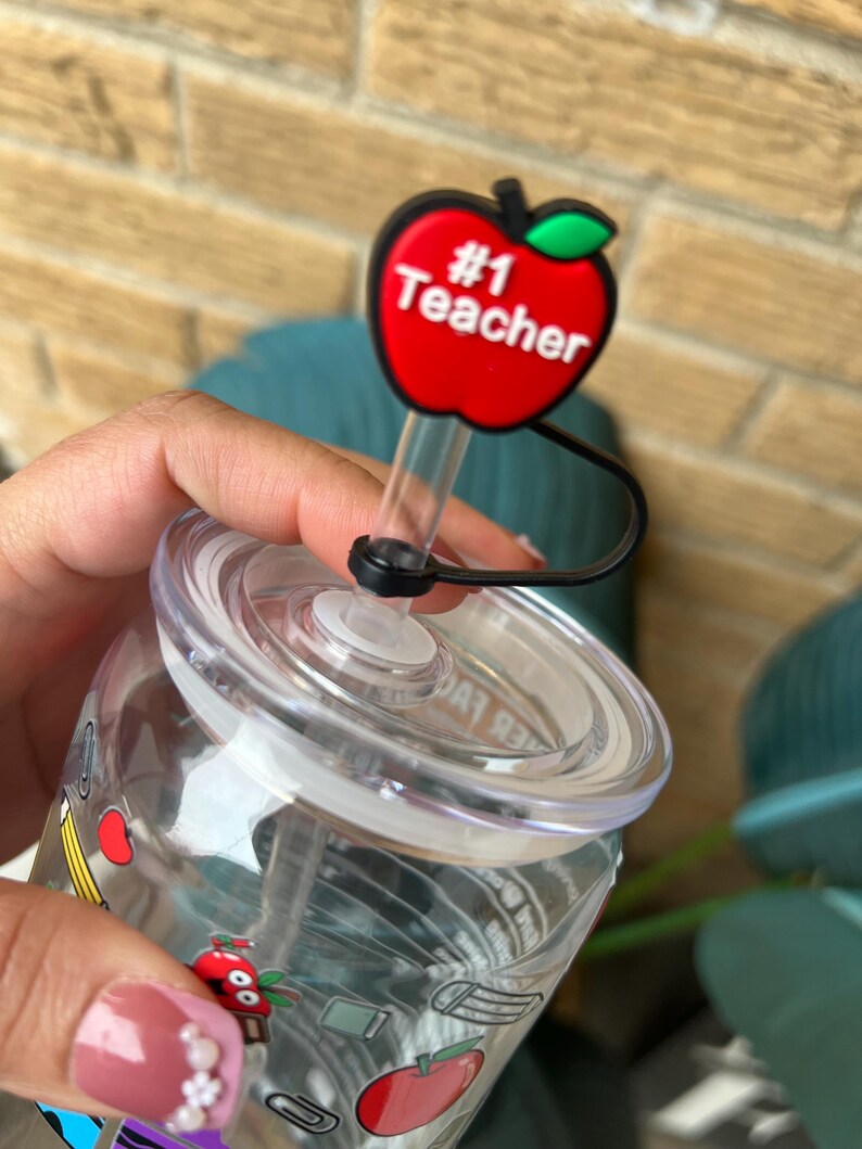 May include: A clear plastic water bottle with a red apple lid that says "#1 Teacher". The bottle has a straw and is decorated with school supplies like pencils, rulers, and apples.