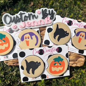 May include: A collection of Halloween-themed car coasters. The coasters feature designs of a jack-o'-lantern, a ghost, and a black cat. The coasters are tan with black, orange, purple, and green accents. The words "Custom by JennB" are displayed.