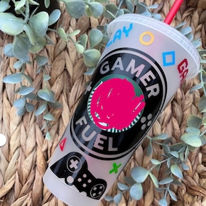 May include: A white reusable cup with a black and green gamer fuel logo. The cup has a black and white video game controller graphic and colorful symbols around the top.