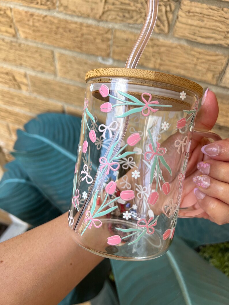 May include: A clear glass mug with a bamboo lid and a pink and green floral design. The mug has a straw and a handle.