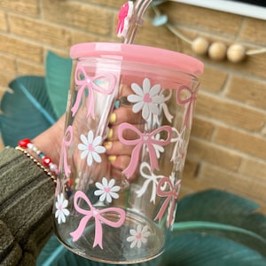 May include: A clear glass tumbler with a pink lid and a pink straw. The tumbler has a pink and white floral design with bows.