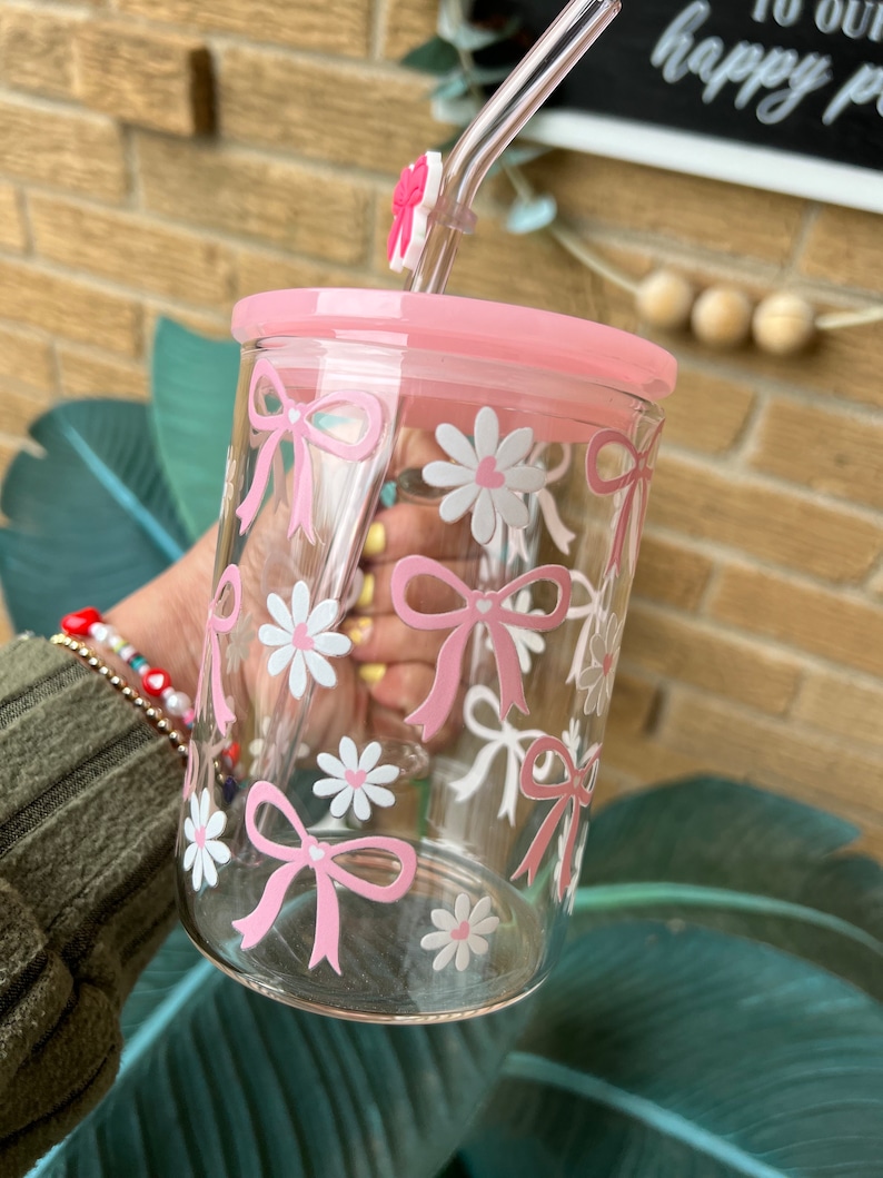 May include: A clear glass tumbler with a pink lid and a pink straw. The tumbler has a pink and white floral design with bows.
