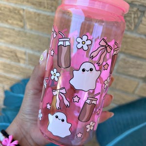 May include: A pink-tinted glass tumbler with a matching lid, adorned with whimsical illustrations. The design includes cute ghost figures, bows, flowers, stars, and iced coffee motifs. The tumbler is held by a person.