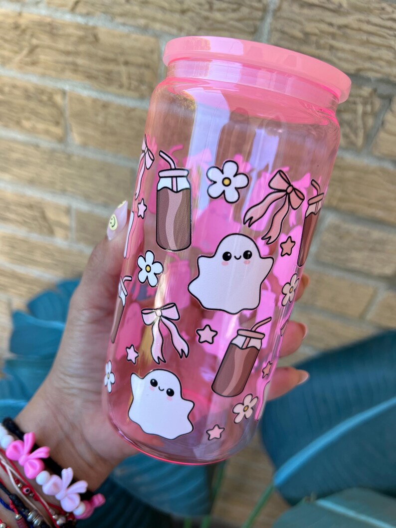 May include: A pink-tinted glass tumbler with a matching lid, adorned with whimsical illustrations. The design includes cute ghost figures, bows, flowers, stars, and iced coffee motifs. The tumbler is held by a person.
