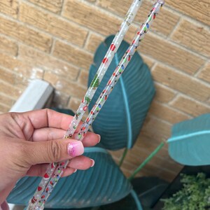 Op de afbeelding: Twee doorzichtige plastic rietjes met kerstthema-ontwerpen. E&eacute;n rietje heeft een kerstboom en sneeuwvlokken, de andere heeft kleurrijke kerstverlichting. De rietjes worden tegen een bakstenen muur en grote groene bladeren gehouden.