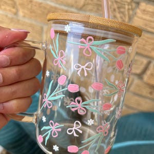 May include: A clear glass mug with a bamboo lid and a straw. The mug features a floral pattern with pink tulips, green leaves, and white flowers.