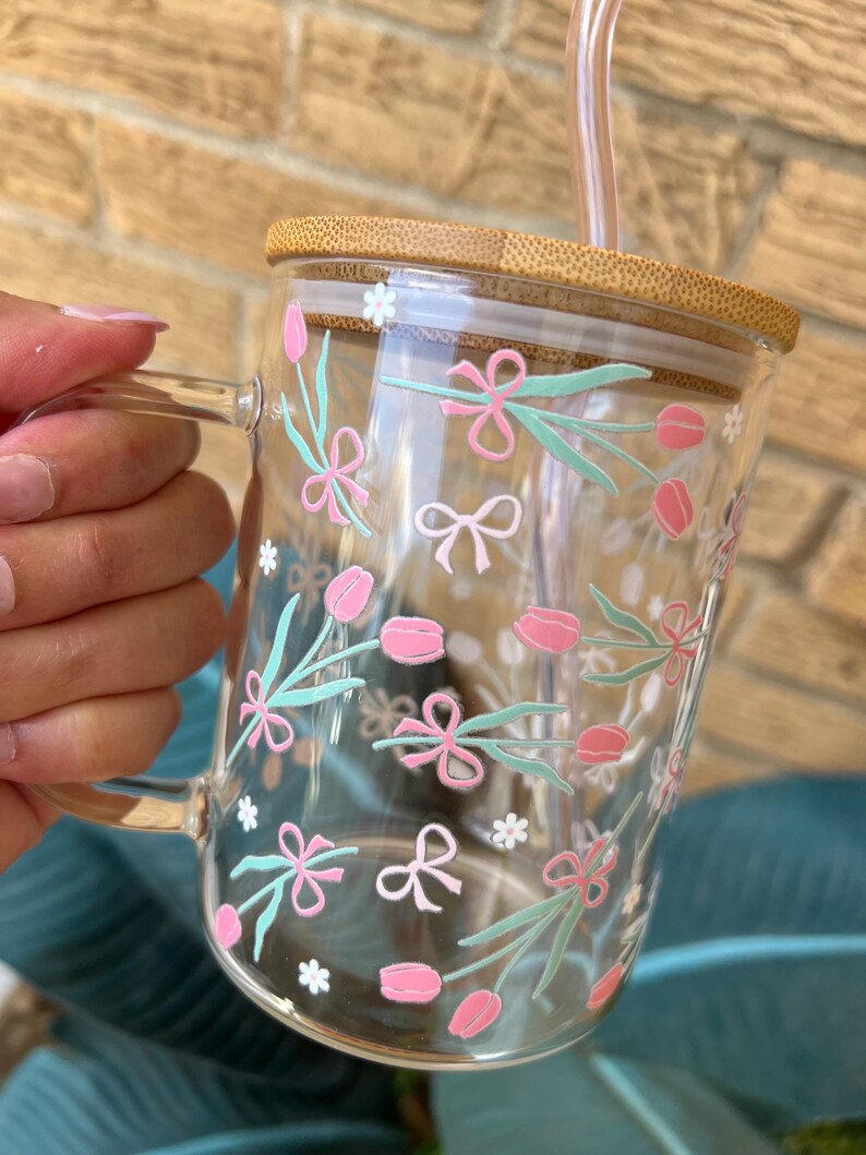 May include: A clear glass mug with a bamboo lid and a straw. The mug features a floral pattern with pink tulips, green leaves, and white flowers.