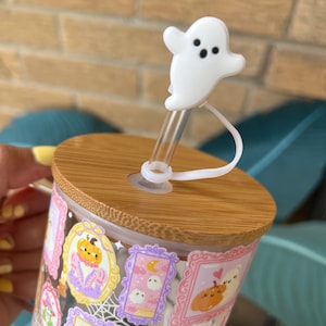 May include: A clear glass tumbler with a bamboo lid and a white ghost-shaped straw topper. The tumbler is decorated with framed Halloween-themed illustrations in pink, purple, and orange. The straw is clear.