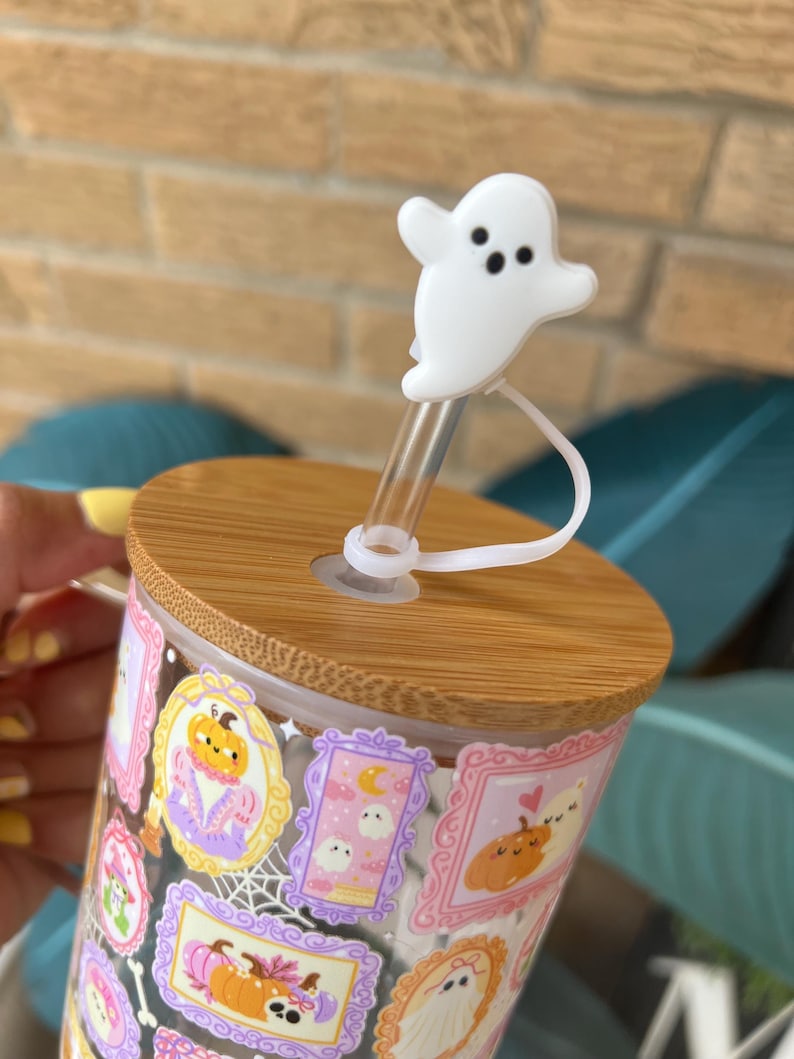 May include: A clear glass tumbler with a bamboo lid and a white ghost-shaped straw topper. The tumbler is decorated with framed Halloween-themed illustrations in pink, purple, and orange. The straw is clear.