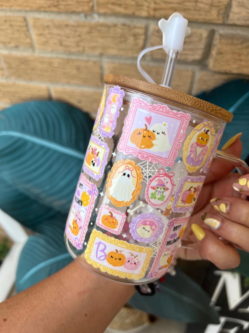 May include: A clear glass mug with a bamboo lid and straw, decorated with Halloween-themed illustrations in ornate frames. The mug features pumpkins, ghosts, and the word "Boo". The straw has a white flower-shaped stopper.