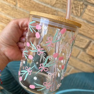 May include: A clear glass tumbler with a bamboo lid and a reusable straw. The tumbler features a floral pattern with pink flowers, green leaves, and white bows.