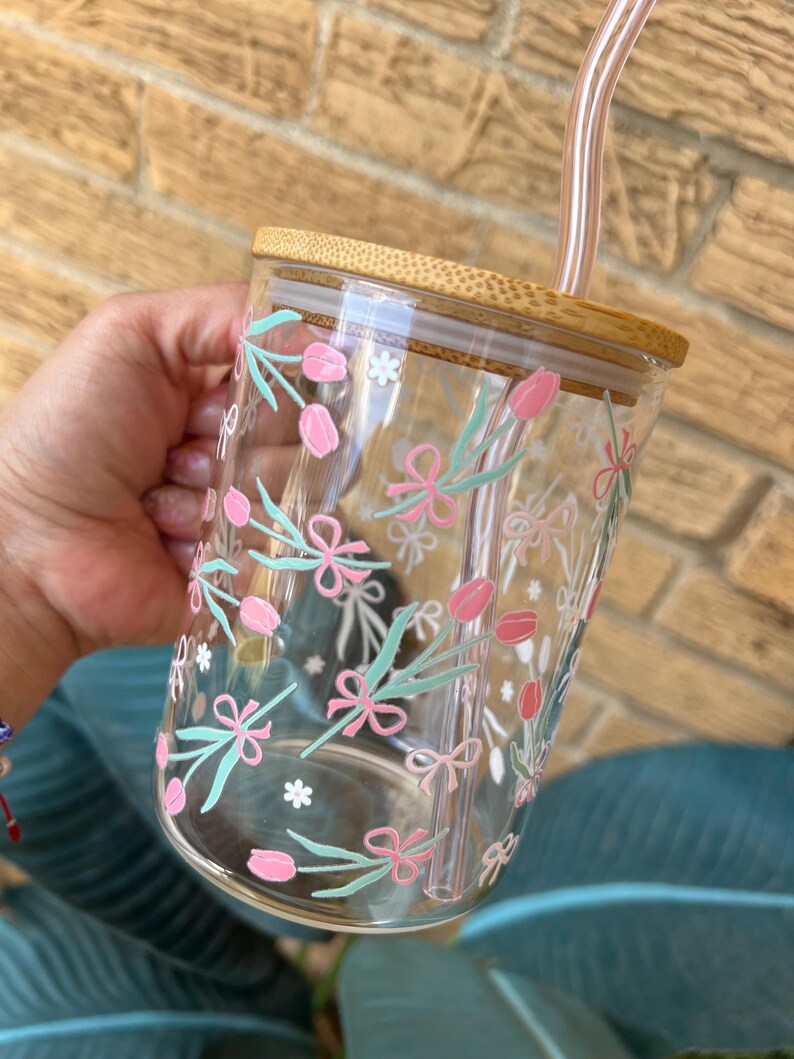 May include: A clear glass tumbler with a bamboo lid and a reusable straw. The tumbler features a floral pattern with pink flowers, green leaves, and white bows.