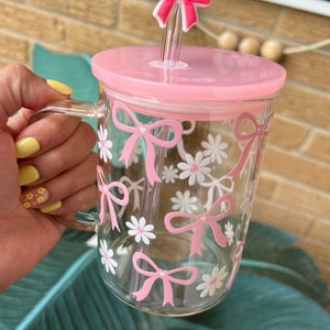 May include: A clear glass mug with a pink lid and a pink straw. The mug has a pink bow and white daisy pattern.