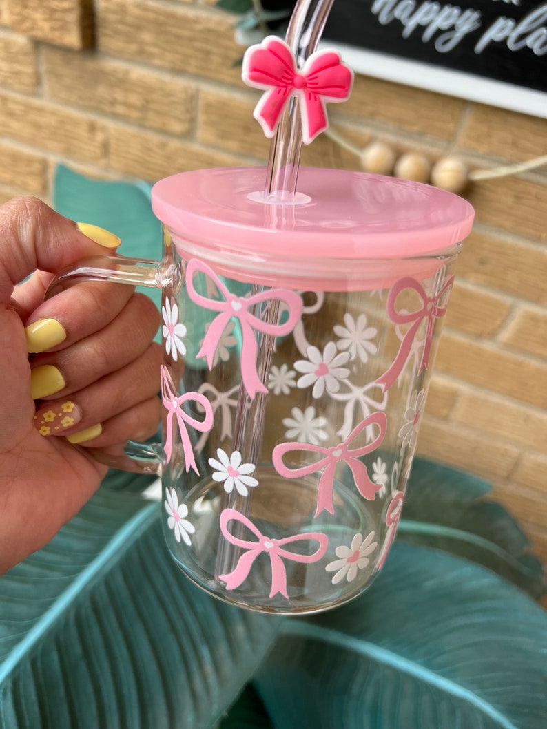 May include: A clear glass mug with a pink lid and a pink straw. The mug has a pink bow and white daisy pattern.