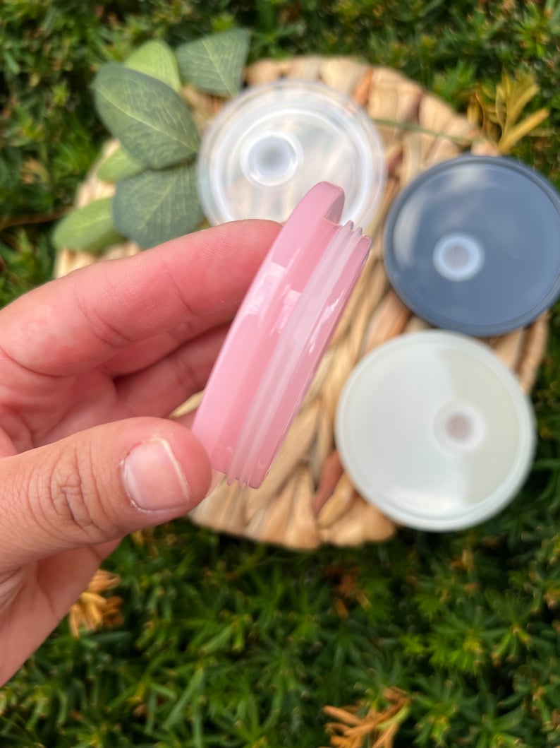 May include: A close-up of a pink, clear plastic lid with a white ring around the edge. The lid is being held by a hand.