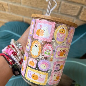 May include: A clear glass tumbler with a bamboo lid and a white ghost-shaped straw topper. The glass is decorated with framed Halloween-themed illustrations of pumpkins, ghosts, and other festive elements. The words "Happy Halloween" are visible.