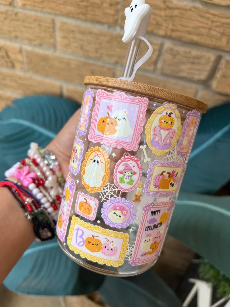 May include: A clear glass tumbler with a bamboo lid and a white ghost-shaped straw topper. The glass is decorated with framed Halloween-themed illustrations of pumpkins, ghosts, and other festive elements. The words "Happy Halloween" are visible.