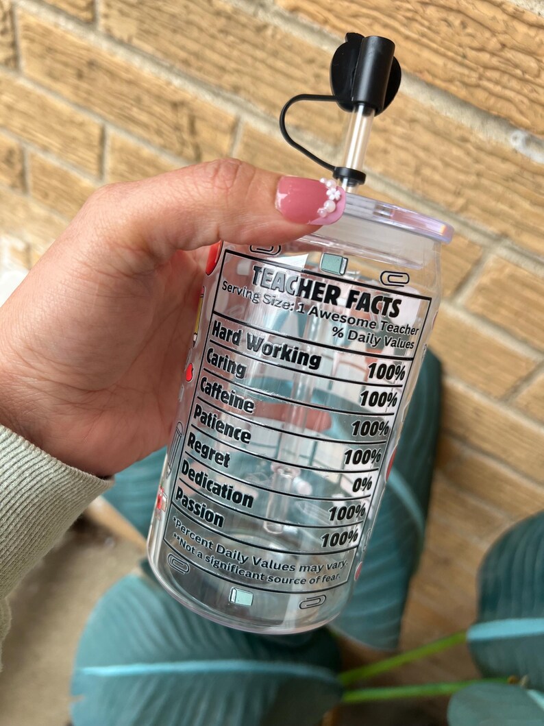 May include: A clear plastic tumbler with a black lid and straw. The tumbler has a label that reads "Teacher Facts" and lists various qualities with percentages, such as "Hard Working 100%", "Caring 100%", "Caffeine 100%", "Patience 100%", "Regret 0%", "Dedication 100%", and "Passion 100%".