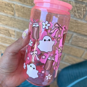 May include: A clear glass tumbler with a pink lid, adorned with cute illustrations. The design includes white ghosts, flowers, bows, and coffee drinks. The tumbler is held in a hand.