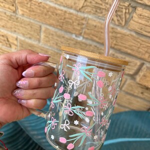 May include: A clear glass mug with a bamboo lid and a pink and green floral design. The mug has a handle and comes with a clear plastic straw.