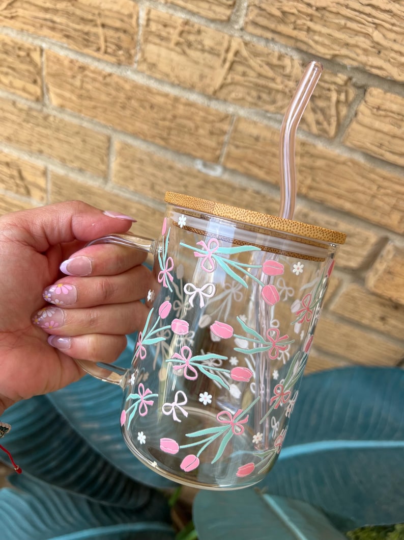 May include: A clear glass mug with a bamboo lid and a pink and green floral design. The mug has a handle and comes with a clear plastic straw.
