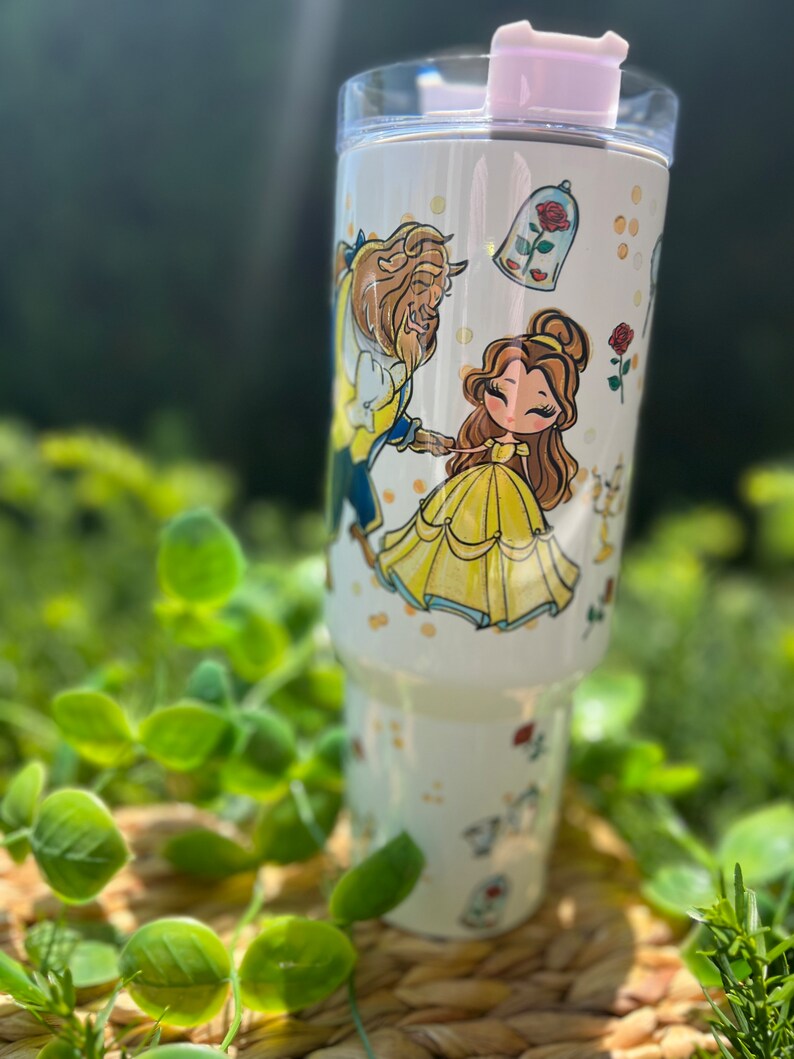 Op de afbeelding: Een witte beker met illustraties van Belle en het Beest uit Belle en het Beest. Het ontwerp bevat de personages, een roos en een glazen koepel. De beker heeft een lichtroze deksel en is omgeven door groen.