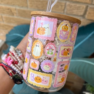 May include: A clear glass tumbler with a bamboo lid and a white straw. The tumbler is decorated with framed Halloween-themed illustrations of pumpkins, ghosts, and other spooky elements in pink, orange, and purple. The words "Happy Halloween" are also visible.