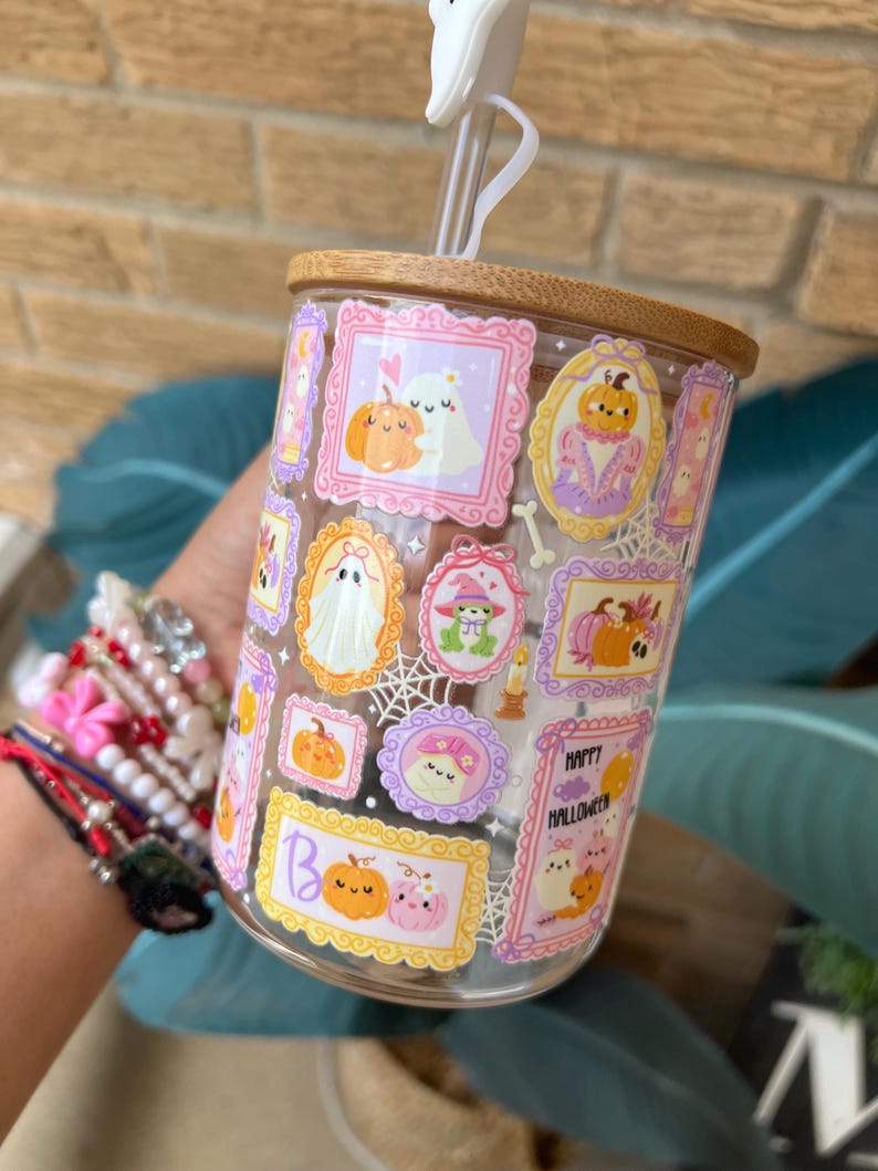 May include: A clear glass tumbler with a bamboo lid and a white straw. The tumbler is decorated with framed Halloween-themed illustrations of pumpkins, ghosts, and other spooky elements in pink, orange, and purple. The words "Happy Halloween" are also visible.