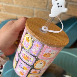 May include: A clear glass tumbler with a wooden lid and a white ghost-shaped straw topper. The tumbler is decorated with framed Halloween-themed illustrations in various colors, including pumpkins, ghosts, and bats.