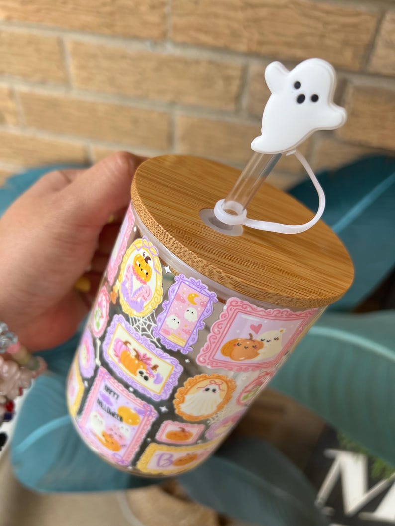 May include: A clear glass tumbler with a wooden lid and a white ghost-shaped straw topper. The tumbler is decorated with framed Halloween-themed illustrations in various colors, including pumpkins, ghosts, and bats.