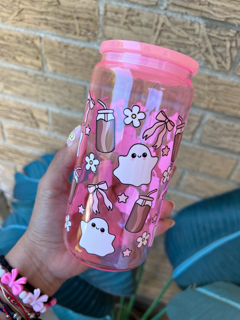 May include: A pink-tinted glass tumbler with a matching lid, decorated with cartoon ghosts, bows, flowers, and drink illustrations. The tumbler is designed for cold beverages.
