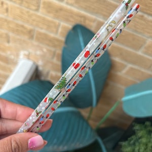 Op de afbeelding: Twee doorzichtige plastic eetstokjes met kerstthema's. E&eacute;n eetstokje heeft een patroon van kerstverlichting, een kerstboom en een rode want. De andere eetstokje heeft een patroon van hulst en sneeuwvlokken.