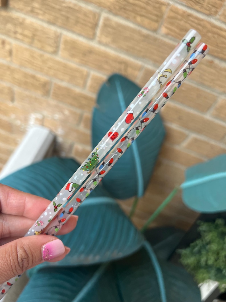 Op de afbeelding: Twee doorzichtige plastic eetstokjes met kerstthema's. E&eacute;n eetstokje heeft een patroon van kerstverlichting, een kerstboom en een rode want. De andere eetstokje heeft een patroon van hulst en sneeuwvlokken.
