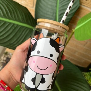 May include: A clear glass reusable water bottle with a bamboo lid and a black and white cartoon cow design. The bottle has a black and white striped straw.