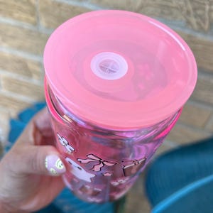 May include: A clear tumbler with a pink lid and a white straw. The tumbler is decorated with pink and white cartoon graphics. The lid is a solid pink color. The tumbler is held in a hand.