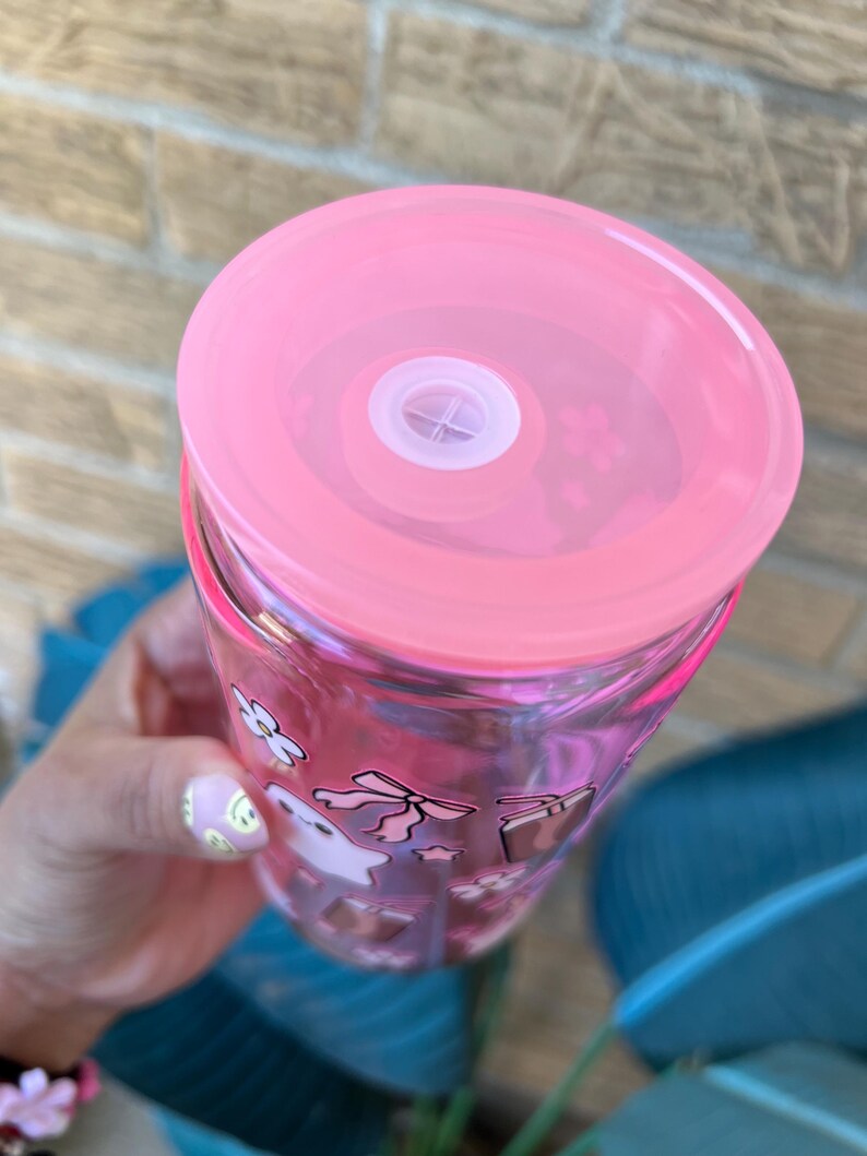 May include: A clear tumbler with a pink lid and a white straw. The tumbler is decorated with pink and white cartoon graphics. The lid is a solid pink color. The tumbler is held in a hand.