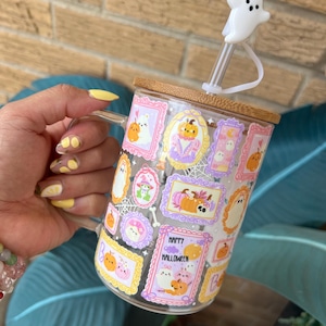 May include: A clear glass mug with a bamboo lid and a white ghost-shaped straw topper. The mug is decorated with Halloween-themed illustrations in frames, including pumpkins and ghosts. The words "Happy Halloween" are visible on the mug.